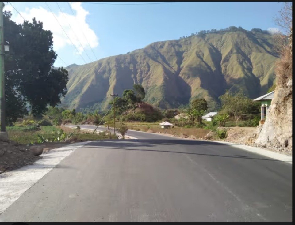 Paved mountain road winding through a lush valley under a bright blue sky with scenic peaks.