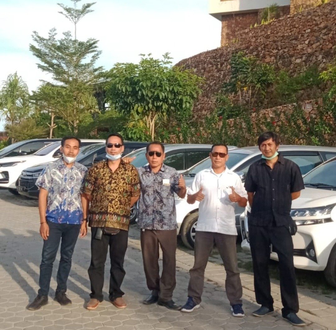A group of five men standing in front of parked cars outdoors wearing batik shirts and sunglasses.